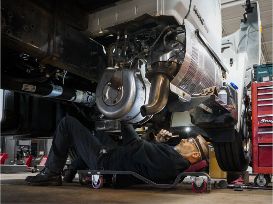 Mechanic lying on a creeper and inspecting the underside of a truck in a repair shop.