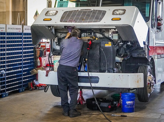Mechanic working under the raised hood of a large delivery truck inside an auto repair shop.